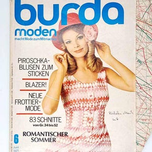 Puede incluir: Una portada vintage de la revista Burda Moden de junio de 1971. La portada presenta a una mujer con un sombrero rojo y un vestido estampado. El título de la revista está en letras azules grandes. El texto incluye "PIROSCHKA-BLUSEN ZUM STICKEN" y "ROMANTISCHER SOMMER".