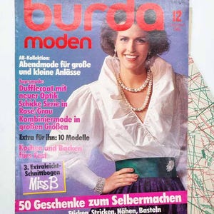Può includere: Una copertina di rivista vintage con una donna che indossa una camicetta bianca e una collana di perle. Il titolo della rivista "Burda Moden" è in lettere rosse. Il testo include "Abendmode" e "50 Geschenke zum Selbermachen". La copertina ha uno sfondo blu.