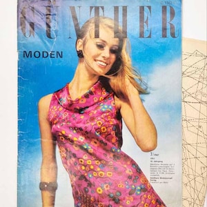 Può includere: Una copertina di rivista vintage Gunther Moden del luglio 1967 che mostra una persona che indossa un abito rosa senza maniche con un colorato motivo floreale, scollo ad anello e un braccialetto scuro, su sfondo blu con testo nero.