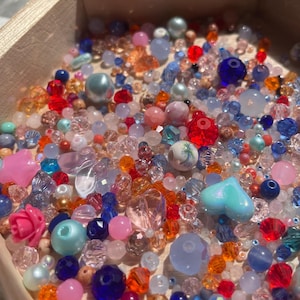 May include: A wooden box overflowing with a variety of colorful beads. The beads are in shades of pink, blue, orange, red, and clear. There are different shapes and sizes, including round, heart-shaped, and faceted beads. A mix of materials, including glass and plastic.