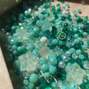 May include: A collection of green and teal beads in various shapes and sizes. The assortment includes faceted beads, star-shaped beads, and a small rose-shaped bead. Some beads are clear, while others are opaque. The beads are scattered in a wooden container.