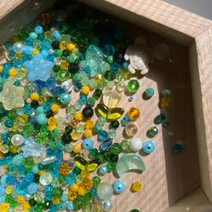 May include: A wooden tray filled with an assortment of colorful beads in shades of blue, green, yellow, and clear. The beads include various shapes like stars, flowers, and a small turtle figurine. The arrangement creates a vibrant, ocean-themed aesthetic.