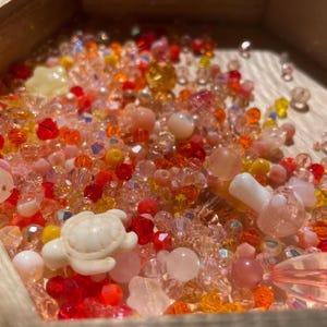 May include: A wooden box filled with a variety of colorful beads and charms. The assortment includes clear, red, orange, yellow, and pink beads, along with small turtle and mushroom charms. The beads are of various shapes and sizes, creating a textured and vibrant display.