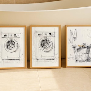 Op de afbeelding: Drie ingelijste kunstdrukken met schetsen van wasgoedgerelateerde items. Twee prints tonen wasmachines, terwijl de derde een wasmand met kleding en handdoeken laat zien. De frames zijn lichtbruin.