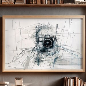 Peut inclure: Impression artistique abstraite représentant un croquis d'un appareil photo et d'une personne. L'œuvre est réalisée dans des tons de gris et de noir, avec l'accent mis sur l'objectif de l'appareil photo. L'impression est encadrée dans un cadre en bois clair.