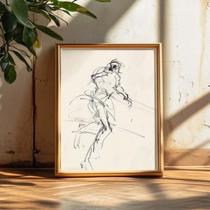May include: A framed black and white sketch of a figure in motion. The artwork is in a gold frame, set against a textured wall and wooden floor. The drawing style is loose and expressive, with visible lines.