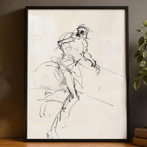 May include: Black framed artwork featuring a sketch of a person in motion. The artwork is a black and white line drawing on a cream-colored background. The frame is black and the artwork is displayed on a wooden surface.