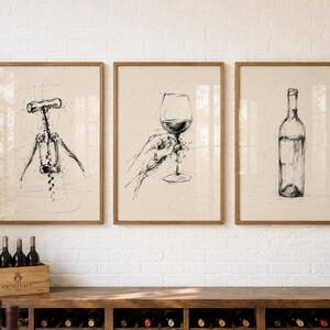 May include: Three framed sketches of wine-related items. The artwork includes a corkscrew, a hand holding a wine glass, and a wine bottle. The sketches are in black ink on a cream-coloured background, with wooden frames. The artwork is displayed above a wooden wine rack.