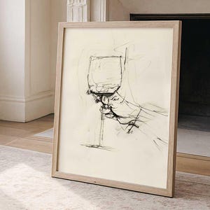 Può includere: Un'opera d'arte incorniciata con un'illustrazione di un bicchiere di vino tenuto da una mano. Il disegno è in bianco e nero, con uno sfondo beige chiaro. La cornice è in legno chiaro, aggiungendo un tocco di eleganza.