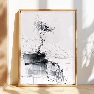 May include: Abstract black and white art print of a tree on a cliff, framed in a natural wood frame. The artwork features a minimalist style with expressive lines and brushstrokes, creating a dramatic landscape scene. The print is displayed on a wooden surface.