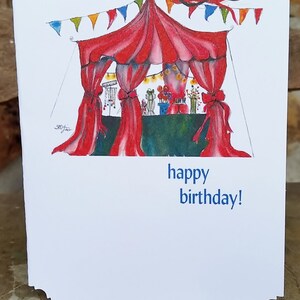 May include: A white birthday card with a watercolor illustration of a circus tent with red curtains and colorful bunting. The text "happy birthday!" is printed in blue.