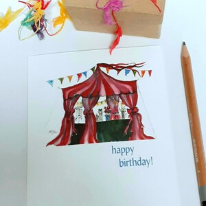 May include: A birthday card with a watercolor illustration of a circus tent in red and white, with colorful bunting. The card has the words "happy birthday!" in blue. A brown gift box with pink and red ribbons and a pencil are also in the image.