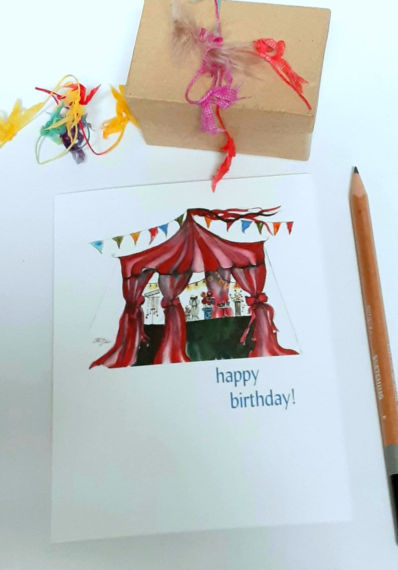 May include: A birthday card with a watercolor illustration of a circus tent in red and white, with colorful bunting. The card has the words "happy birthday!" in blue. A brown gift box with pink and red ribbons and a pencil are also in the image.