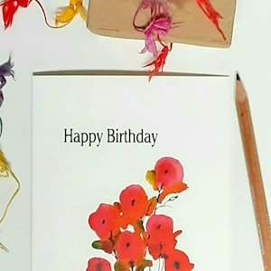 May include: A white greeting card with the text "Happy Birthday" and a watercolor floral design in shades of red and orange. A brown gift box is tied with colorful ribbons. A pencil rests on the side.