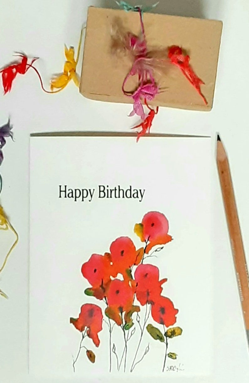 May include: A white greeting card with the text "Happy Birthday" and a watercolor floral design in shades of red and orange. A brown gift box is tied with colorful ribbons. A pencil rests on the side.