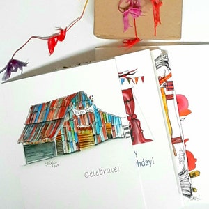 May include: A stack of greeting cards with watercolor illustrations. One card features a colorful barn with the text "Barn Dance." Another says "Celebrate!" and a third says "Happy Birthday!" A small gift box is tied with colorful ribbons.