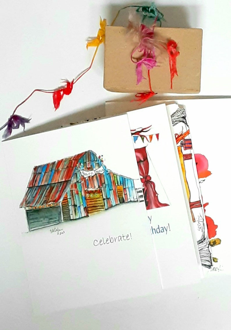May include: A stack of greeting cards with watercolor illustrations. One card features a colorful barn with the text "Barn Dance." Another says "Celebrate!" and a third says "Happy Birthday!" A small gift box is tied with colorful ribbons.