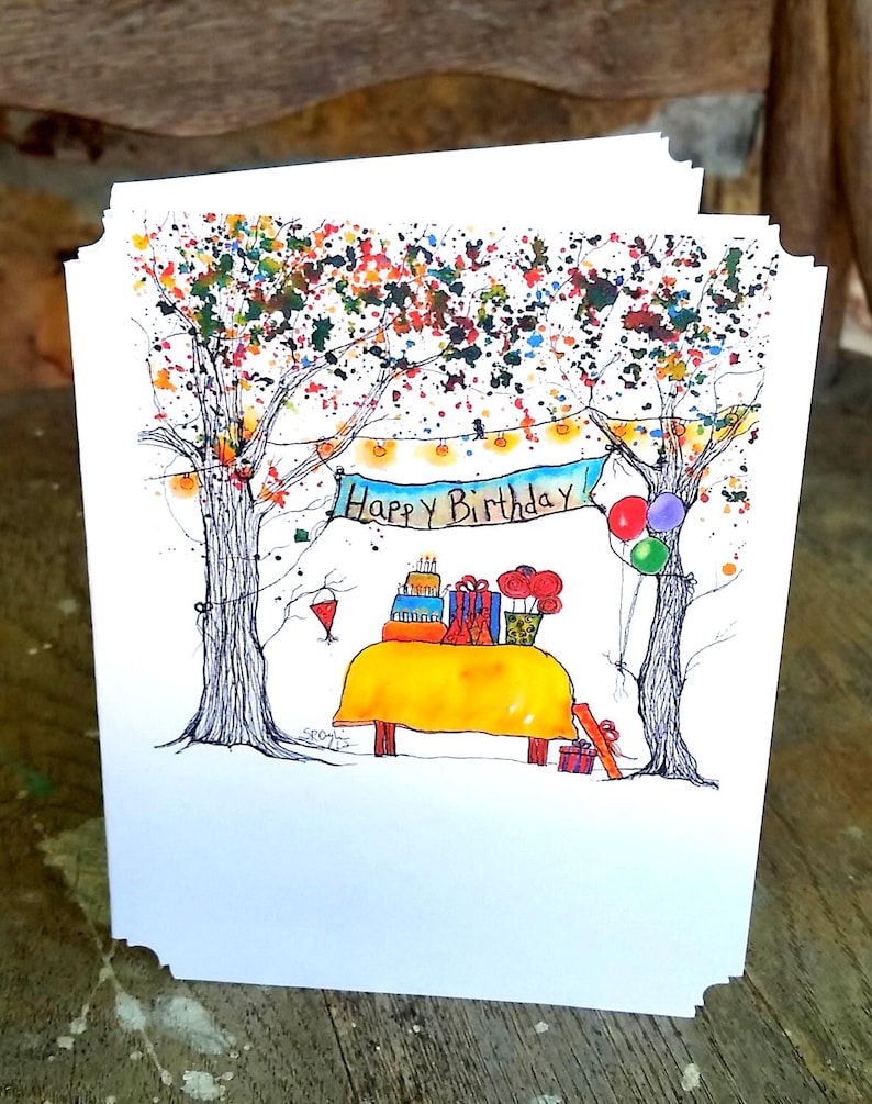 May include: A white greeting card with a hand-drawn illustration of a birthday scene. The artwork features a yellow table with gifts and a cake, set between two trees with colorful foliage. A banner reads "Happy Birthday".