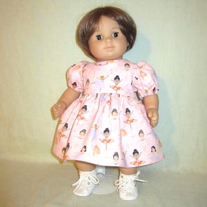 May include: A doll wearing a pink dress with a pattern of ballerinas in various poses. The dress has short, puffed sleeves and a full skirt. The doll is also wearing white shoes with laces.
