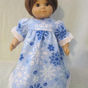May include: A doll with short brown hair wearing a light blue dress featuring a white and dark blue snowflake and paw print pattern. The dress has short puffed sleeves with white lace trim and a matching white lace hem at the bottom edge.