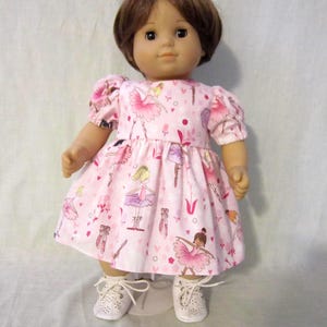 May include: A doll wearing a pink dress with a ballerina print, including ballerinas, ballet shoes, and hearts. The doll has brown hair and is wearing white shoes with laces. The dress has short, puffed sleeves.