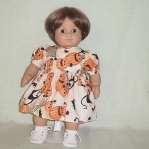 May include: A doll with short brown hair wearing a cream-colored dress featuring an orange pumpkin, black bat, and spiderweb print. The dress has short puffed sleeves and a gathered skirt. The doll wears white lace-up shoes with a perforated design.