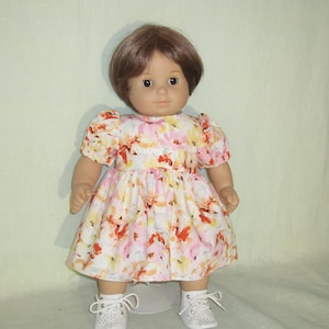 May include: A doll wearing a floral print dress with short sleeves and a gathered skirt. The dress features a pattern of pink, orange, and yellow flowers on a white background. The doll has brown hair and is wearing white shoes.