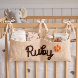 May include: A beige fabric cot organiser with two pockets, featuring the name Ruby in dark brown cursive lettering and a small orange crocheted flower. The organiser is tied to a wooden cot with fabric ribbons and contains baby supplies.