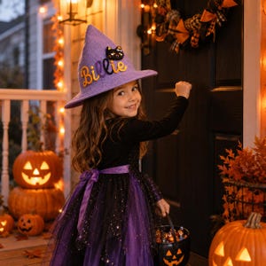 May include: A lavender pointed witch hat featuring the name Billie in yellow cursive lettering and a small black cat patch. The hat is made of a textured fabric and is worn with a black long-sleeved dress with a purple tulle skirt overlay.