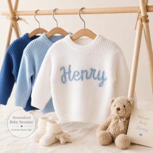May include: Three long-sleeve knit jumpers hanging on wooden hangers. The front jumper is white with the name Henry embroidered in blue yarn. Behind it are light blue and dark blue jumpers. A beige teddy bear and white booties sit below.