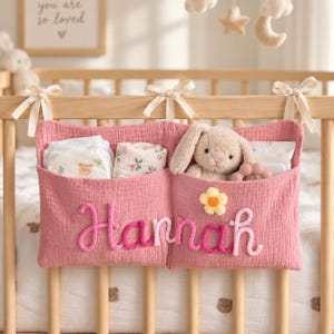 May include: A dusty pink fabric cot organiser with two pockets, tied to a wooden cot with cream ribbons. The name Hannah is embroidered in pink yarn across the front, accented by a small yellow flower. It holds nappies, a cloth, and a plush bunny.