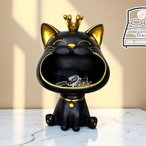 Puede incluir: Contenedor de almacenamiento negro con forma de gato con detalles dorados, que incluyen una corona, orejas y collar. El contenedor tiene una boca abierta para guardar cosas y está sobre una superficie de mármol blanco. El contenedor está lleno de joyas.
