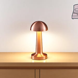 May include: A copper-coloured table lamp with a mushroom-shaped shade. The lamp has a cylindrical base and stem, and it is illuminated. The lamp is on a wooden surface. The image also includes a logo that says "Sleep Sanctuary Design, Bed Accessories."