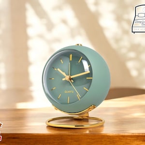 Puede incluir: Un reloj despertador redondo verde con detalles dorados y movimiento de cuarzo. El reloj está sobre una superficie de madera.