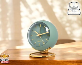 May include: A green round alarm clock with gold accents and a quartz movement. The clock is sitting on a wooden surface.