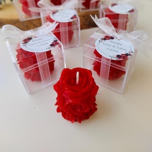 May include: Red rose-shaped candles in clear boxes tied with white ribbons. Each box has a label with text and floral accents. The candles are a vibrant red, and the overall presentation suggests a wedding or event favor.