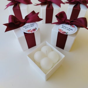 May include: White favor boxes with burgundy ribbon and a floral design with the text "Sydney & Justin June 2, 2024". The boxes contain small white candles.