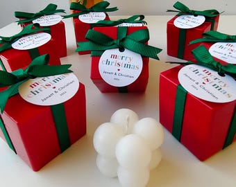 Könnte beinhalten: Rote Geschenkboxen mit grünem Band und einem weißen Kreis mit dem Text "merry christmas" und den Namen "Janett & Christian" und dem Jahr "2024". Im Vordergrund befindet sich eine weiße Blasenkerze.