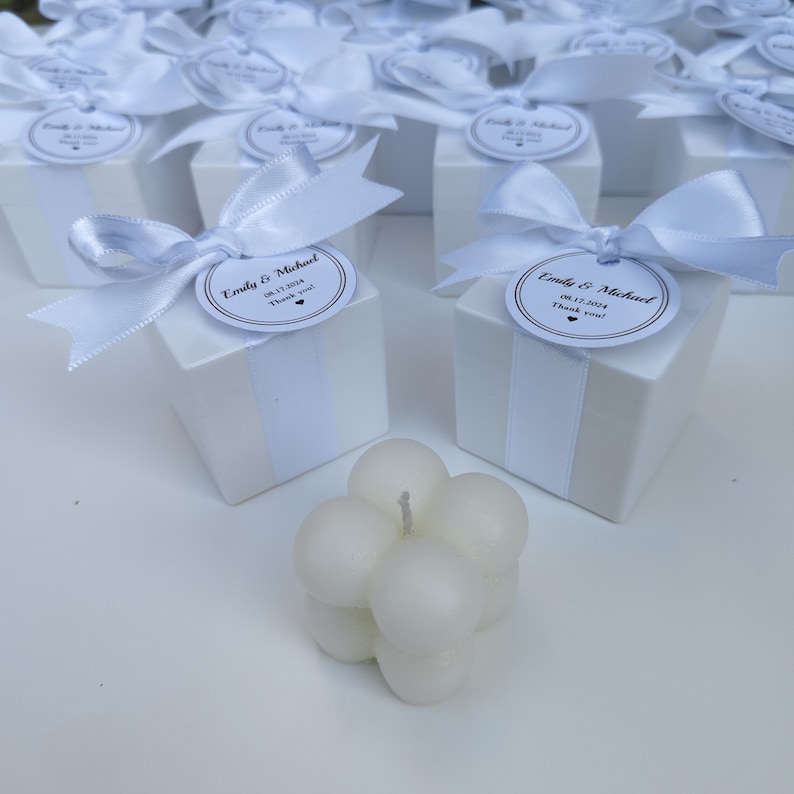 May include: White favor boxes with white satin ribbon and a round white tag with black text that reads "Emily & Michael 08.17.2024 Thank you!"  A white bubble candle is in the foreground.