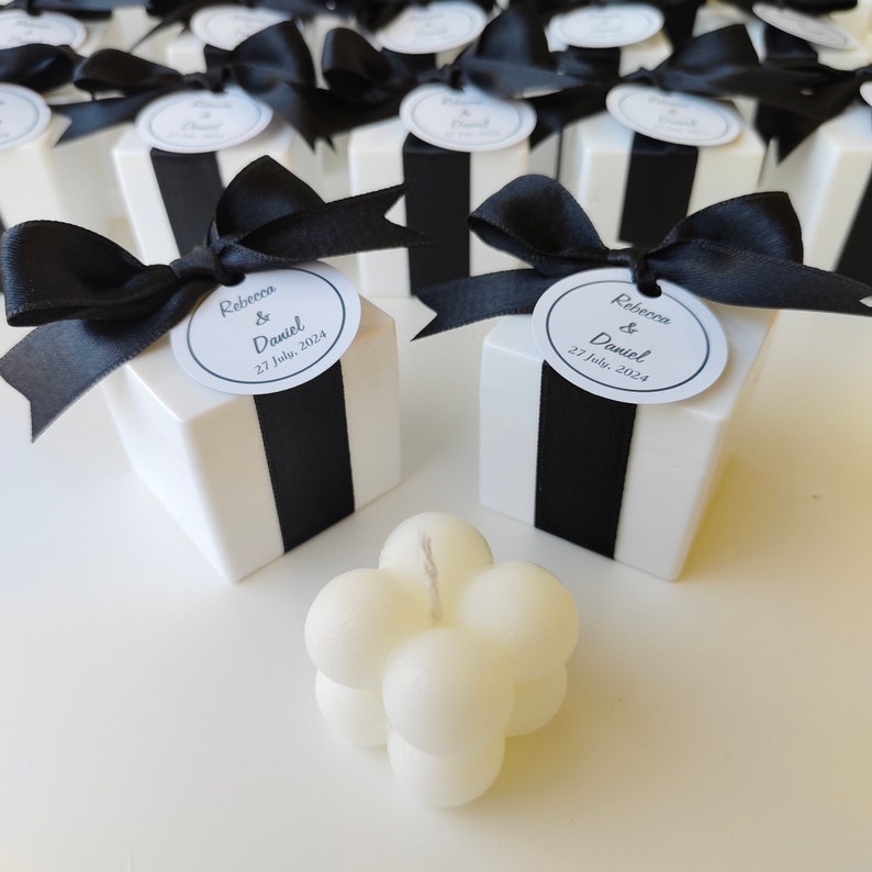 May include: White favor boxes with black ribbon and a circular tag that reads "Rebecca & Daniel 27 July, 2024". A white bubble candle is in the foreground.