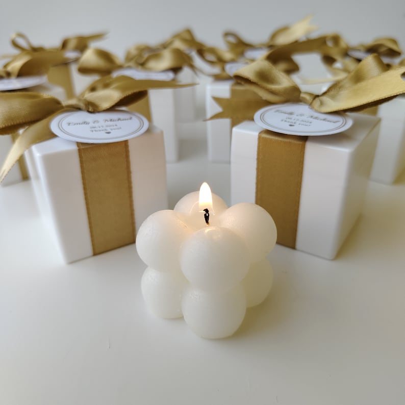 May include: White favor boxes with gold ribbon and a personalized label that says "Emily & Michael" with a wedding date. The boxes are surrounded by white bubble candles with a lit wick.