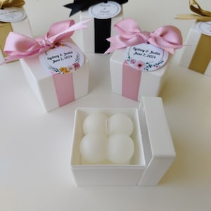 May include: Four white favor candles in a white box with a pink ribbon and a floral wedding favor tag that reads "Sydney & Justin June 2, 2024".