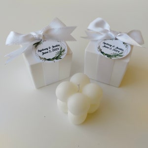 May include: Two white gift boxes with white bows and personalized labels that say "Sydney & Justin June 2, 2024". A white, round, bubble candle is in the center of the image.