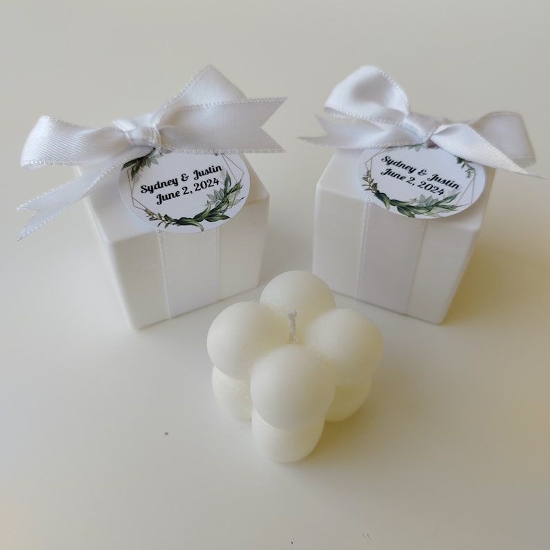 May include: Two white gift boxes with white bows and personalized labels that say "Sydney & Justin June 2, 2024". A white, round, bubble candle is in the center of the image.