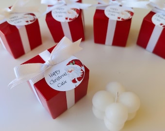 Könnte beinhalten: Fünf kleine rote Geschenkboxen mit weißem Band und einem Weihnachtsmann-Etikett mit der Aufschrift "Frohe Weihnachten Catie". Eine weiße Kerze in Blumenform steht vor den Boxen.