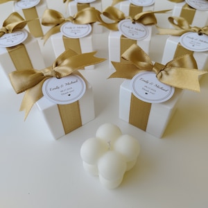 May include: White favor boxes with gold ribbon and a round label that says "Emily & Michael 08.17.2024 Thank you!"  A white candle with a round shape is in the foreground.