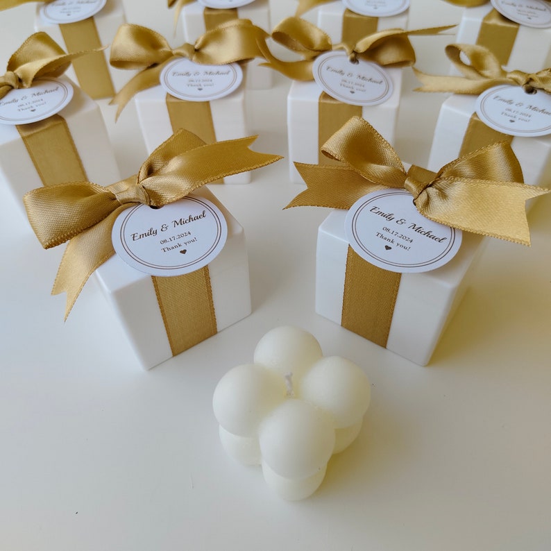 May include: White favor boxes with gold ribbon and a round label that says "Emily & Michael 08.17.2024 Thank you!"  A white candle with a round shape is in the foreground.