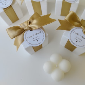 May include: White favor boxes with gold ribbon and personalized labels. The labels read "Emily & Michael" with the date "08.17.2024" and "Thank you!" A white scented candle with a four-petal flower design is in the foreground.