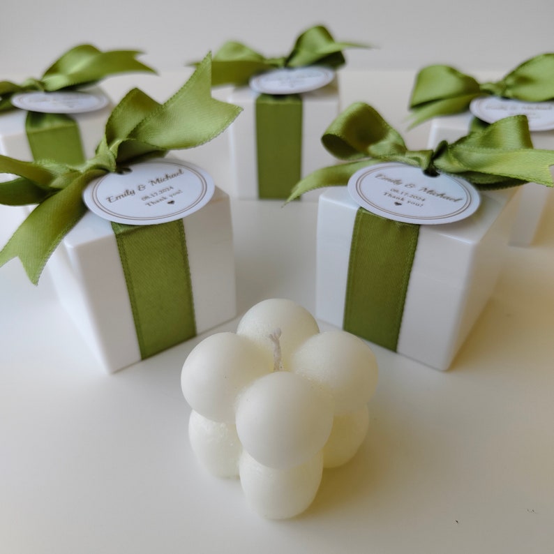 May include: White favor boxes with green ribbon and personalized labels, tied with a bow. The boxes are arranged on a white surface with a white bubble candle in the center.