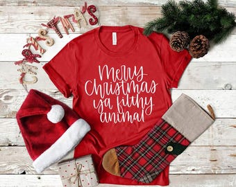 Könnte beinhalten: Rotes T-Shirt mit weißem Text, der "Frohe Weihnachten, du dreckiges Tier" lautet. Das Shirt liegt auf einer weißen Holzoberfläche mit einer roten Weihnachtsmütze, einem rot-schwarzen karierten Strumpf und einem Weihnachtskranz.